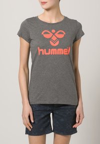 Camiseta gris de manga corta con un destacado logo y diseño gráfico en naranja de "hummel". Combinada con pantalones cortos oscuros de estampado.
