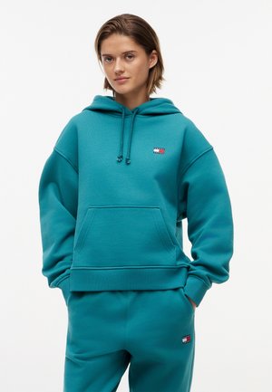 Νεαρός με τιρκουάζ φούτερ με κουκούλα και αντίστοιχο παντελόνι φόρμας με μικρά λογότυπα Tommy Jeans, τα χέρια στις τσέπες, ουδέτερο φόντο.