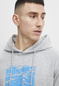 Grå hættetrøje med et frontgrafik, der viser blå og grå bølgemønstre. Den har en snoretrækshætte og er lavet af blødt, textured stof.
