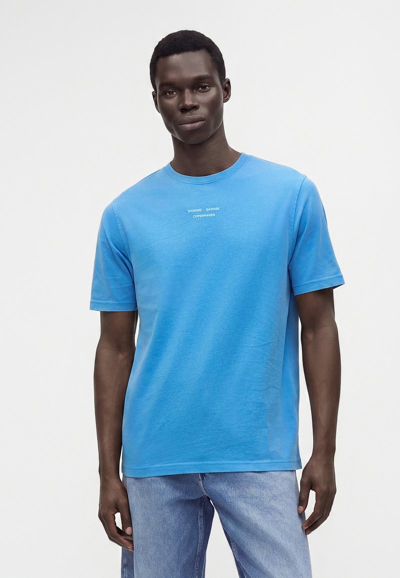 Jeune homme portant un t-shirt bleu vif à manches courtes avec un petit texte blanc et un jean bleu clair, debout devant un fond uni.