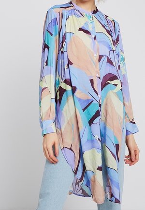 Tunika z długim rękawem zapinana na guziki, z pastelowym abstrakcyjnym kwiatowym wzorem w kolorach niebieskim, fioletowym, beżowym i morskim, noszona na jasnoniebieskich dżinsach.