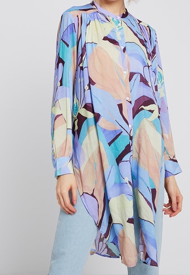 Tunique manches longues boutonnée avec motif floral abstrait pastel en bleu, violet, beige et bleu sarcelle portée sur un jean bleu clair.