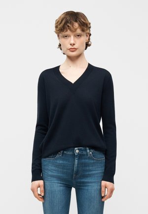 Young person with short curly hair wearing a black V-neck sweater and blue jeans, standing against a plain white background.