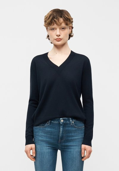 Jeune personne aux cheveux courts et bouclés, portant un pull noir à col en V et un jean bleu, debout devant un fond blanc uni.