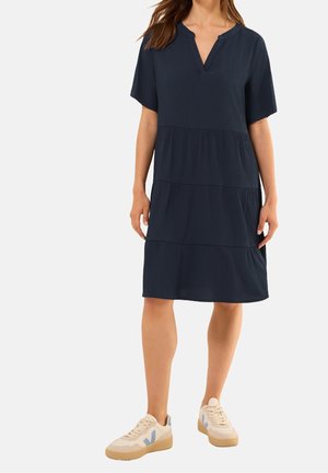 Femme portant une robe ample bleu marine mi-genoux à manches courtes et des baskets beige avec des détails bleus, debout sur un fond uni.