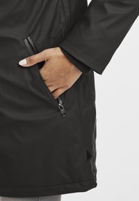 Abrigo negro impermeable con un bolsillo con cremallera en un lado, textura suave y detalle de puño elástico en la manga.