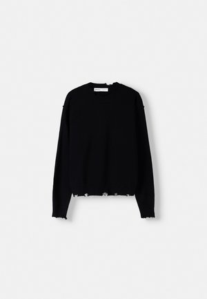 Person iført en sort, slidt striktrøje med frynset kanter og blå jeans, hænderne i lommerne, beskåret ved nakke og talje.
