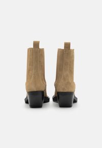 Botines de ante beige con un tacón cilíndrico negro y tiradores en la parte trasera. Textura suave con detalles de costura a lo largo de los lados.