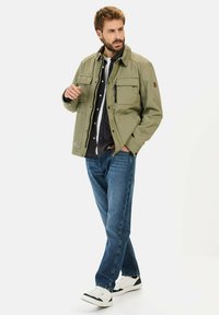 Oliven grøn jakke med flere foran lommer, parret med en mørk button-up skjorte og blå jeans. Model iført hvide sneakers.