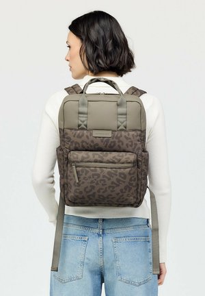 Woman with short dark hair wearing light sweater and jeans, carrying brown leopard-print backpack with front zipper pocket.