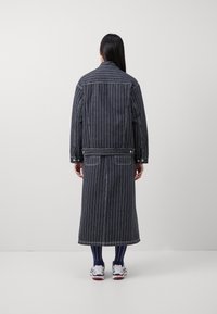 Lang, donker denimjack met witte verticale strepen, voorzien van een aansluitende schouderpas, drukknoopsluiting en een lengte tot onder de knie.