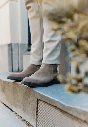 Botas de ante marrón hasta el tobillo con paneles laterales elásticos y suelas de goma negra, combinadas con pantalones de color crema, colocadas sobre un escalón de piedra.