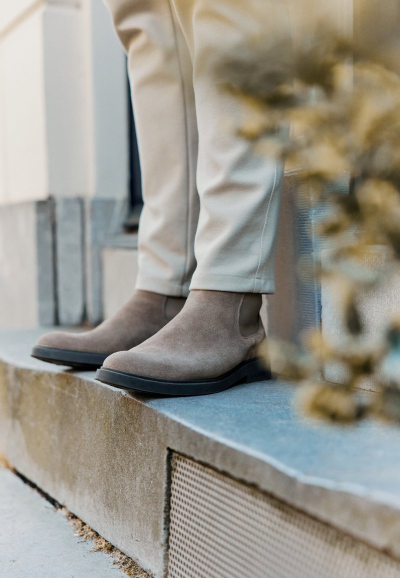 Botas de ante marrón hasta el tobillo con paneles laterales elásticos y suelas de goma negra, combinadas con pantalones de color crema, colocadas sobre un escalón de piedra.