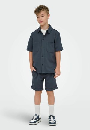 Gutt iført en mørkeblå kortermet skjorte med knapper, matchende shorts med oppbrettede ben, hvite sokker og marineblå sneakers, stående med hendene i lomma.