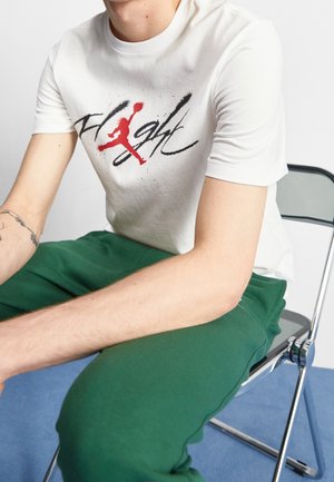 Persona vestida con una camiseta blanca con un gráfico rojo y negro, sentada en una silla de metal con pantalones verdes, frente a un fondo liso.