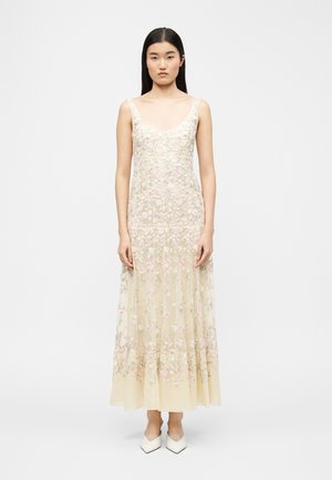 Nő ujjatlan, padlót érő, áttetsző bézs ruhában, rózsaszín virágmotívumos hímzéssel, karjait lazán tartva, fehér hegyes orrú cipőben áll.