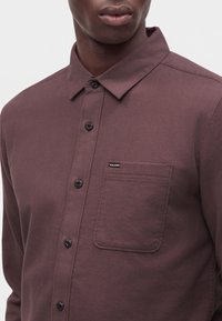 Gros plan d'une personne portant une chemise marron foncé boutonnée avec une poche poitrine comportant une petite étiquette Volcom.
