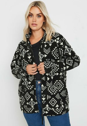 Fleece-Jacke mit einem schwarz-weißen geometrischen Muster, mit einem Reißverschluss vorne, hohem Kragen und zwei Fronttaschen. Weiche Textur.