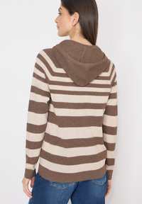 Femme aux cheveux brun foncé portant un sweat à capuche en tricot rayé marron et beige et un jean bleu, tournée de dos sur un fond blanc.