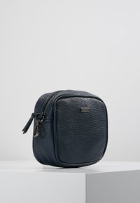 Tmavě modrá kožená kosmetická taštička s texturovaným povrchem. Čtvercový tvar, horní uzávěr na zip a malý stříbrný kovový doplněk.