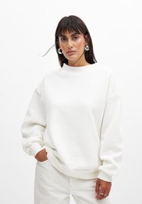 Sudadera blanca con un cuello alto, hombros caídos y puños acanalados. Textura suave y corte relajado, combinada con pantalones color crema.