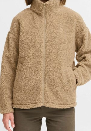 Beige fleecejakke med fuld lynlås, høj krave, struktureret overflade, sidelommer, båret over olivengrønne bukser, hånd i højre lomme.