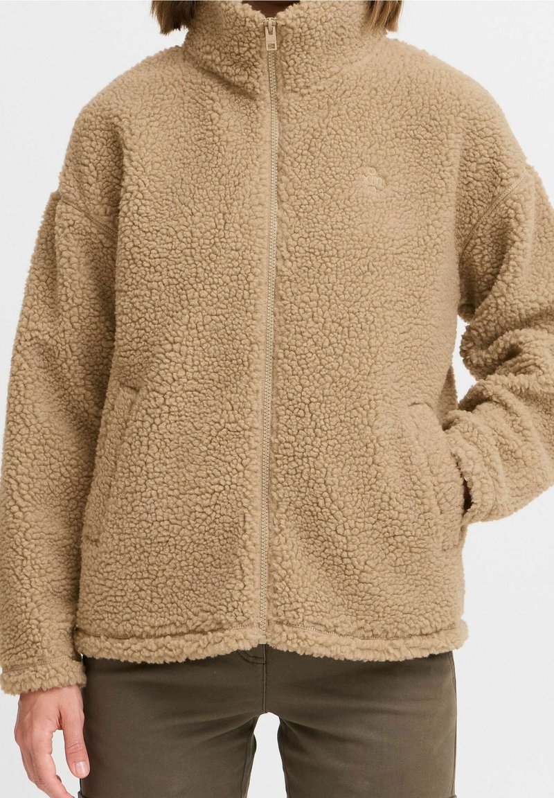 Veste polaire beige à fermeture éclair intégrale, col montant, surface texturée, poches latérales, portée sur un pantalon olive, main dans la poche droite.