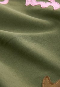 Tissu vert olive à texture lisse, avec des lettres brodées en rose et un patch cousu marron, créant un contraste dans le design.