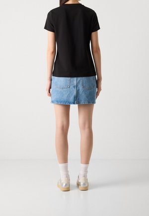 Femme debout de dos, portant un t-shirt noir, une mini-jupe en jean bleu, des chaussettes blanches et des baskets argentées sur fond blanc.