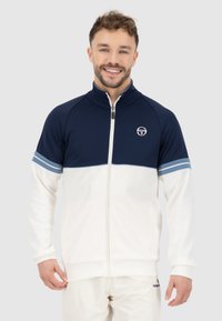 Veste zippée bleu marine et blanche avec un col haut, présentant des rayures horizontales bleues sur les manches et un logo sur la poitrine. Tissu doux.