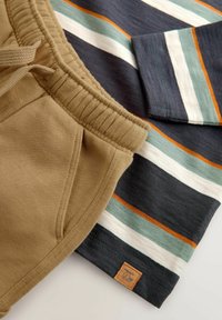 Beige bomulds-joggers med elastisk talje og sidelomme, sammen med en langærmet skjorte med mørke striber, der har teal, hvid og orange detaljer.