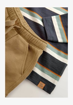 Beige bomulds-joggers med elastisk talje og sidelomme, sammen med en langærmet skjorte med mørke striber, der har teal, hvid og orange detaljer.