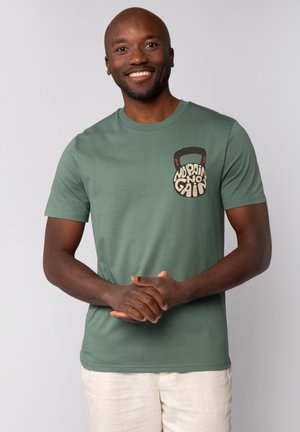 Groene katoenen T-shirt met een vooraanzicht van een kettlebell en de tekst "No Pain No Gain" in crème en zwart. Korte mouwen, ronde hals.