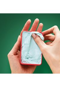 Ljusblå kräm i en röd rektangulär behållare, applicerad med fingrar. En strukturerad, slät konsistens syns inuti behållaren.