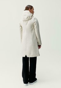 Dlhý krémový kabát s kapucňou, so zošitými švami, rovnou siluetou a dĺžkou po polovicu stehna. Nosený cez čierne nohavice a topánky.