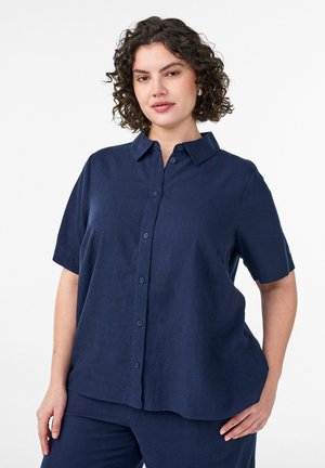 Kortsustega tumelõuendist sinist värvi lühikeste varrukatega nööpidega pluusi ja sobivate pükstega naine seisab ühe käega puusas valgel taustal.