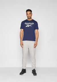 Navy Reebok t-shirt with white logo, gray sweatpants, and black athletic shoes. Model stands upright against a light gray background.