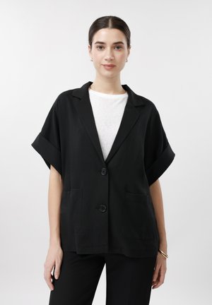 Žena v uvoľnenom čiernom krátkom blejzri s krátkym rukávom cez biele tričko, stojaca na jednoduchom svetlom pozadí.