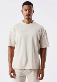 Camiseta oversized beige con cuello redondo y mangas cortas. Presenta un logo "PXP" en tono sobre tono en la parte delantera. Tejido suave y texturizado.