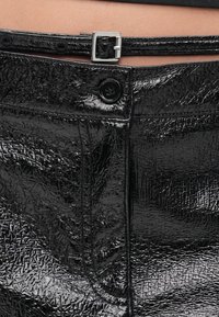 Juodi, blizgūs viniliniai kelnės su tekstūruota paviršiaus struktūra, puoštos sidabriniu sagties diržu ir sagų užsegimu. Artimas juosmens ir detalių vaizdas.