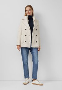 Femme portant un manteau beige à double boutonnage, un col roulé noir, un jean bleu et des baskets blanches, se tenant contre un mur plain.