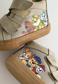 Beige Kinderschuhe mit Klettverschlüssen. Die Seiten zeigen bunte Cartoon-Hundefiguren, das Material ist strukturiert und die Sohle besteht aus braunem Gummi.