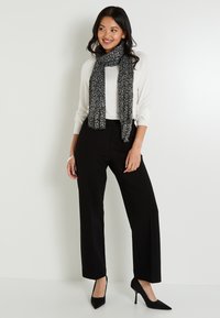 Mujer de pie con una blusa blanca de manga larga, bufanda negra con estampado, pantalones de pierna ancha negros y tacones altos negros, sonriendo y mirando de lado.