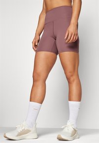 Vysoké fialové sportovní šortky s hladkým materiálem, s těsným střihem a plochými švy, doplněné o bílé ponožky a světlé tenisky.