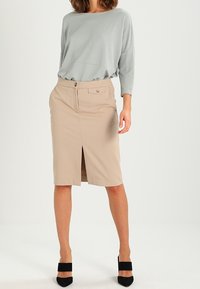 Femme portant un haut à manches longues gris clair, une jupe crayon beige longueur genoux avec fente à l'avant, et des escarpins noirs à bout pointu avec des brides larges.