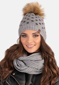 Grå strikket hue med en fluffy brun pom-pom på toppen, præget af mørke prikker og et brunt accentmærke. Kombiner med et gråt struktureret tørklæde.