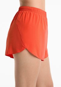 Shorts de sport rouges avec une taille élastique et un ourlet arrondi portés sur la cuisse d'une personne contre un fond blanc.