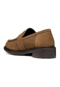 Zapato tipo slip-on de ante marrón con punta redonda, correa decorativa y un tacón de goma texturizado bajo. Vista lateral que destaca la forma y el material del zapato.
