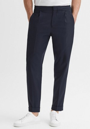 Marineblauwe op maat gemaakte broek met een gladde textuur, geplooide voorkant en licht opgerolde boorden, gecombineerd met witte sneakers.