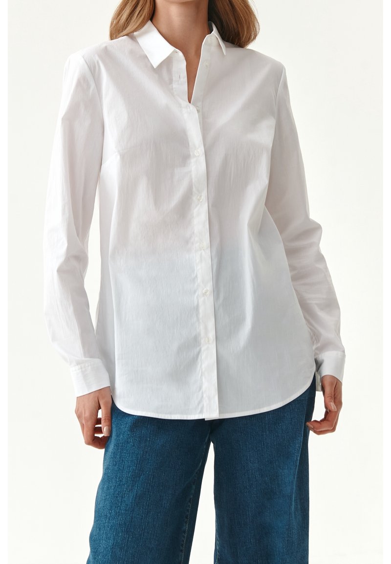 Chemise blanche à manches longues avec col et boutons, fabriquée en coton lisse, présentant une coupe décontractée et une texture douce. Portée avec un jean bleu.